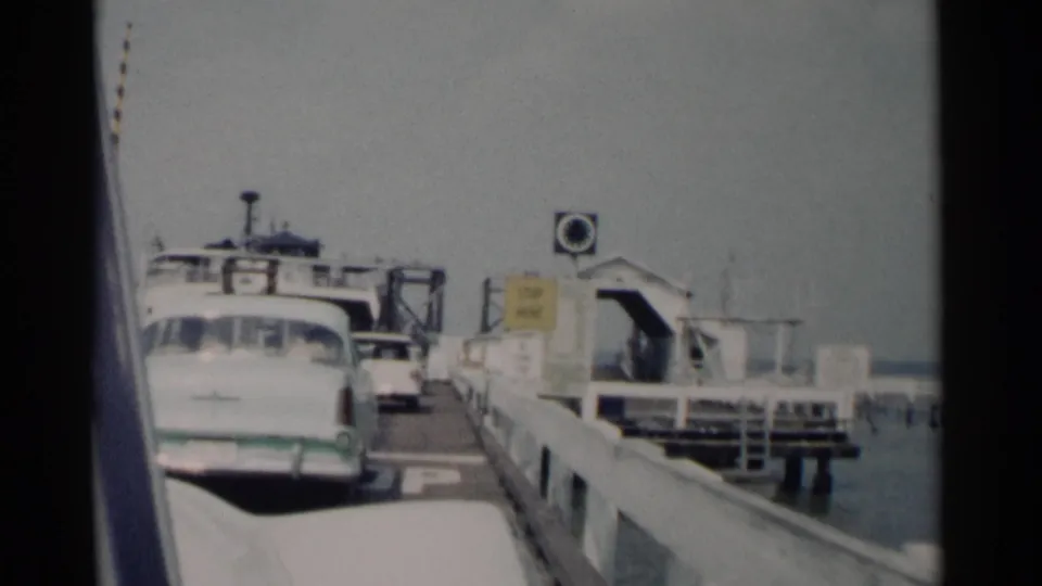 A ferry ramp, dock railings, a white sedan, signage, water, and distant people appear in this 1962 Jamestown, Virginia travel frame.