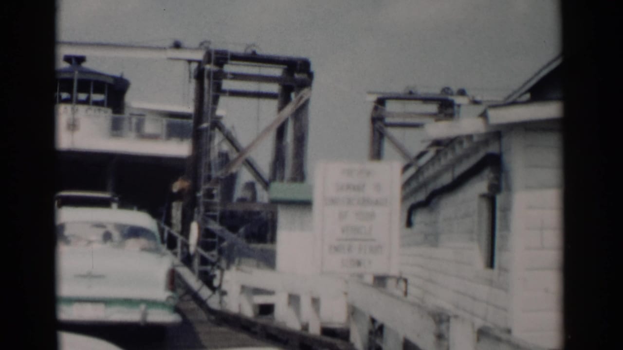White sedan beside a wooden ferry ramp with warning sign and pier machinery.