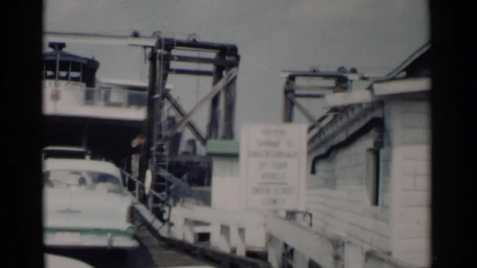 A 1962 Jamestown, Virginia ferry approach shows a white mid-century sedan near a wooden ramp, warning sign, and metal loading structure.
