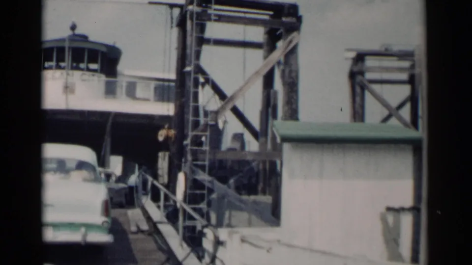 A white mid-century sedan moves through the Jamestown ferry entrance, framed by a booth, ladders, railings, and industrial loading equipment.