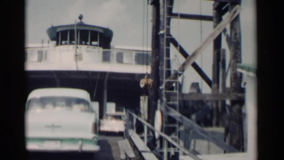 Cars queue at the Jamestown, Virginia ferry ramp, with the white sedan in front, another vehicle behind it, and the control booth overhead.