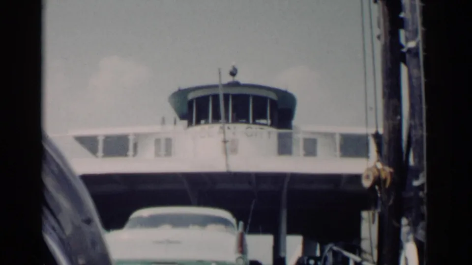 A white early-1960s car moves into the ferry loading area, framed by ramp structure and the pilot house above.