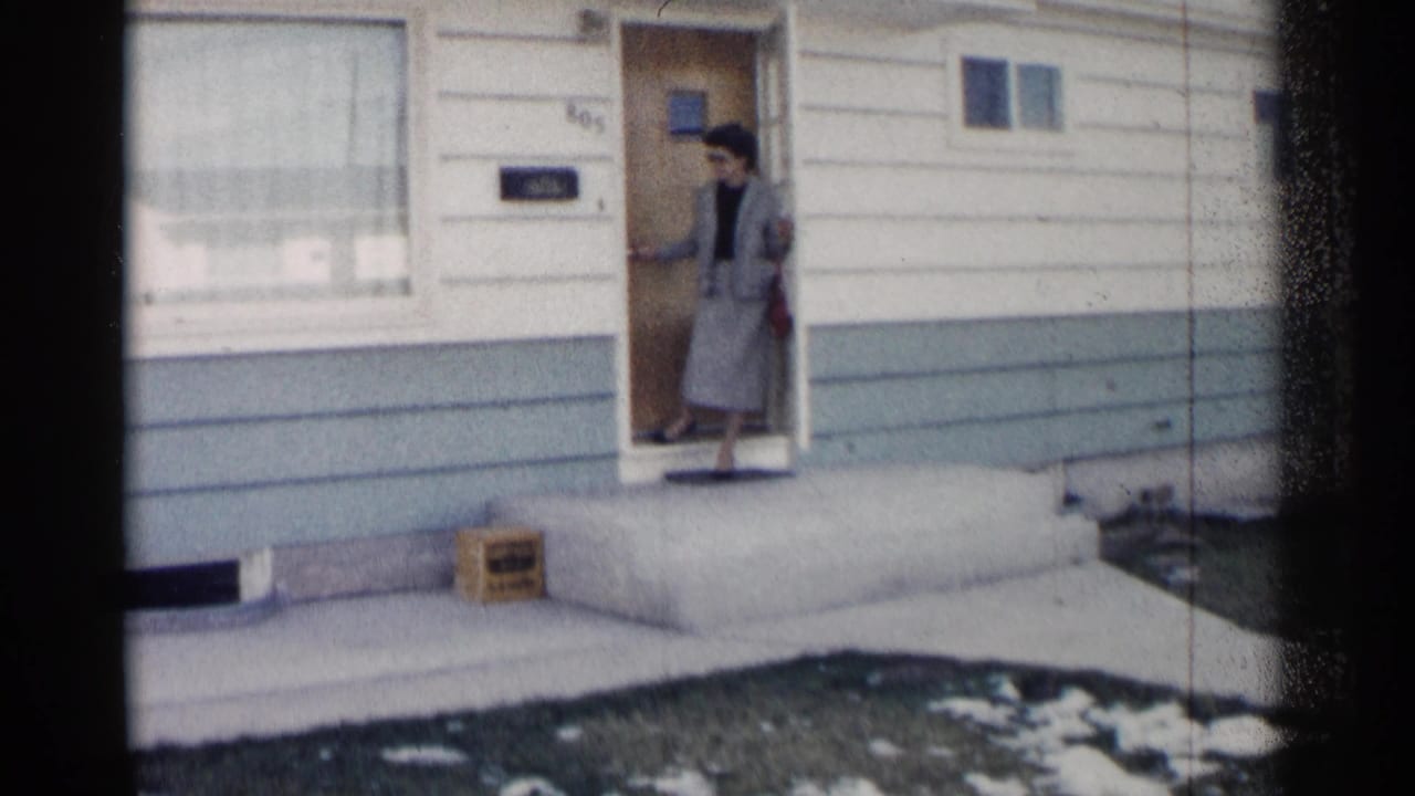 Woman in a gray suit stands in the doorway of a house with siding and porch steps.