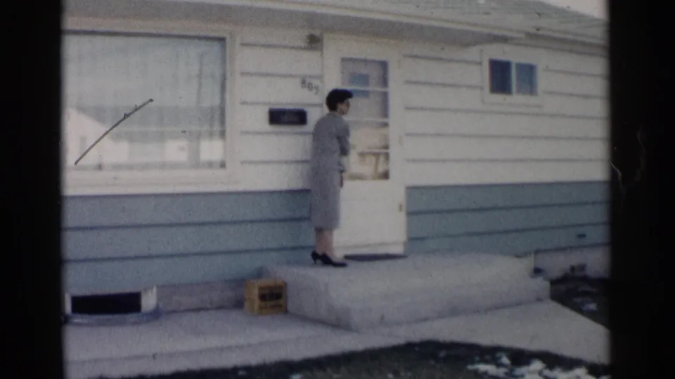 The woman turns toward the front door, appearing to close it on a small porch outside the sided house.