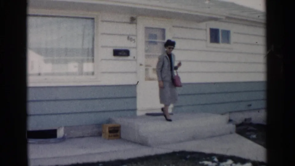A woman in sunglasses carries a handbag while walking away from the closed front door in the 1962 residential scene.