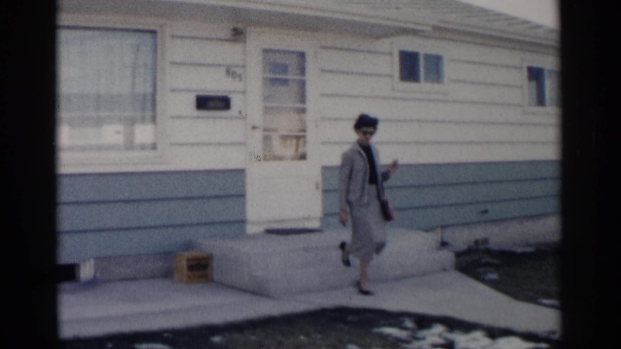 Woman in a gray suit walking away from a white-sided house with blue trim.