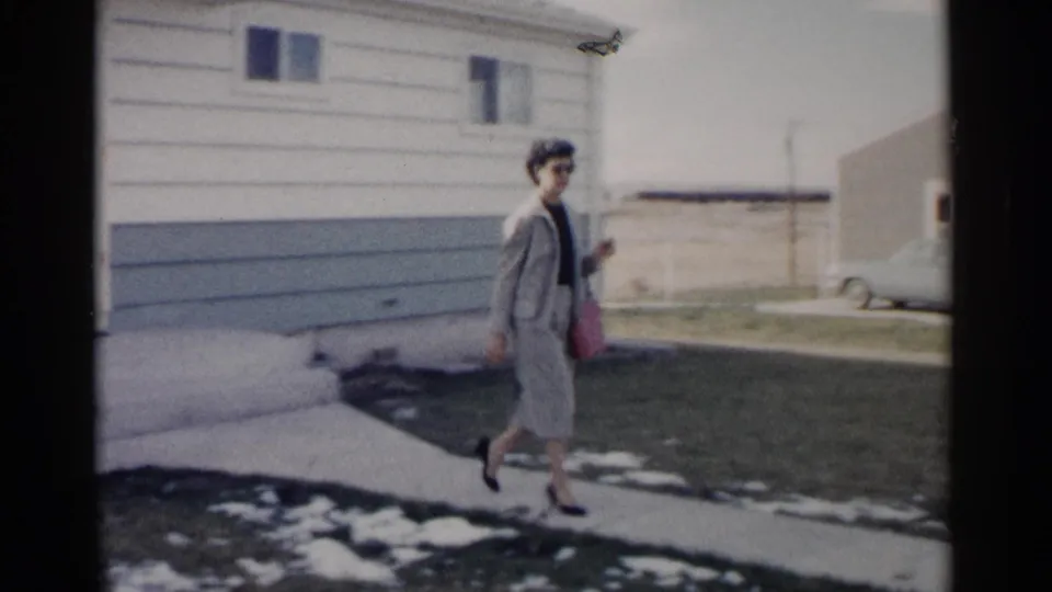 The woman continues along a suburban sidewalk in Washington, DC, with low houses, a lawn, and remaining snow visible behind her.