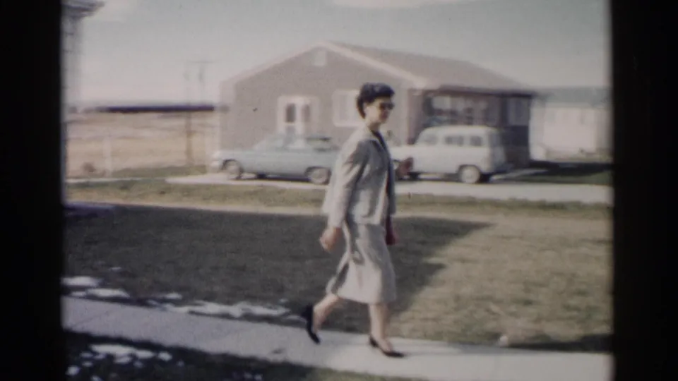 A woman in a gray suit walks past a suburban lawn, with mid-century parked cars and neighboring houses visible in the background.