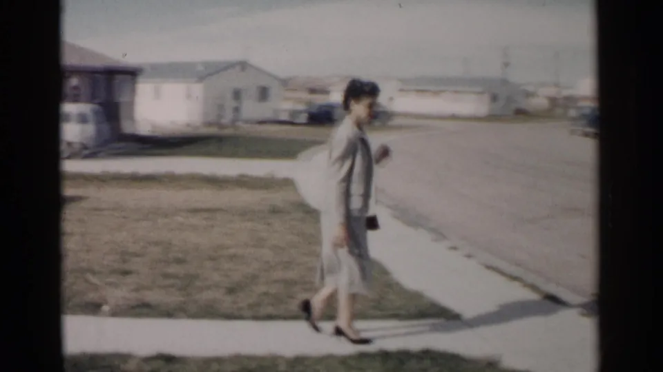 Near the end of the clip, the woman moves along the sidewalk beside an open residential street with low houses in the distance.