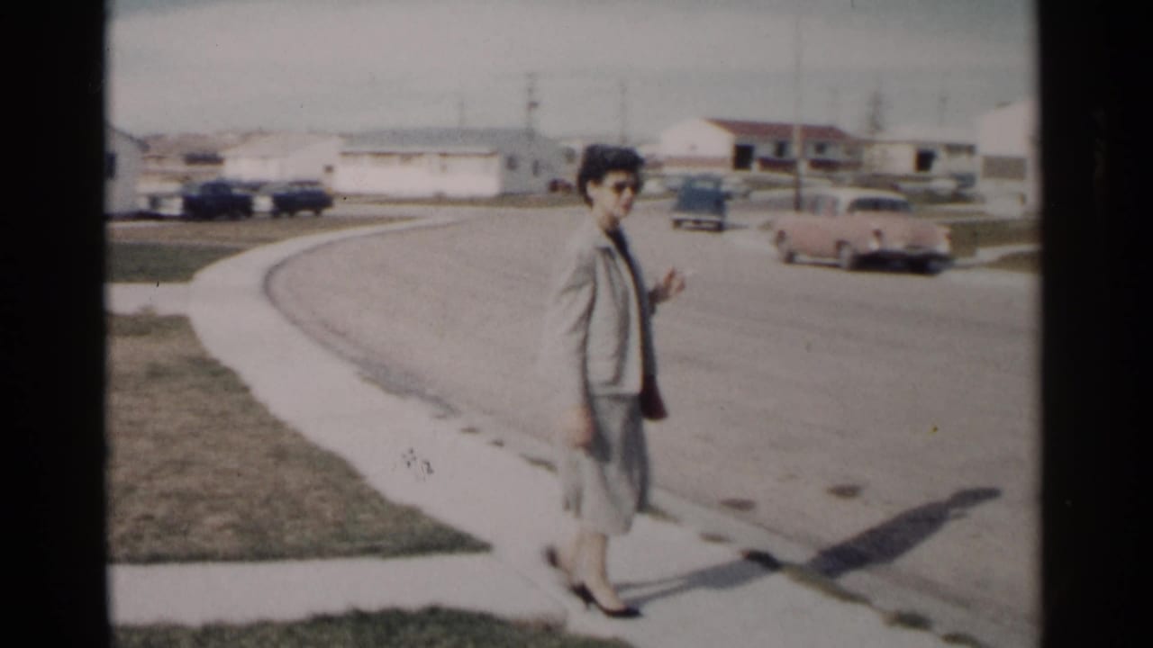 Woman in a light suit walks along a suburban street with houses and parked cars behind her.