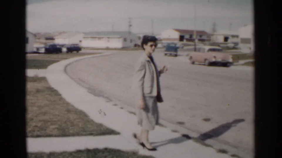 Adult woman in a pale suit and sunglasses walks along a Washington DC suburban street in 1962, with single-story homes and cars in view.
