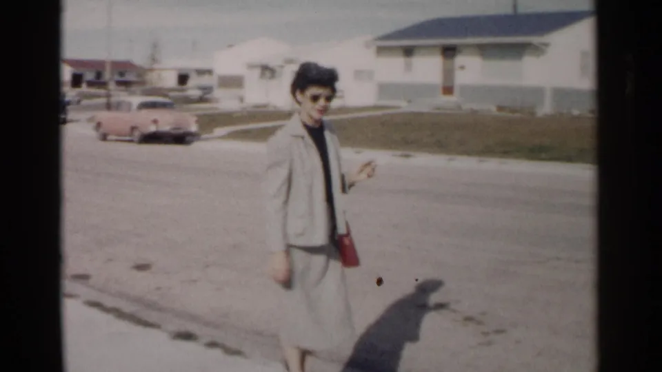 Woman in a light-colored suit carries a red purse while walking through a 1962 Washington DC residential street with low houses behind her.