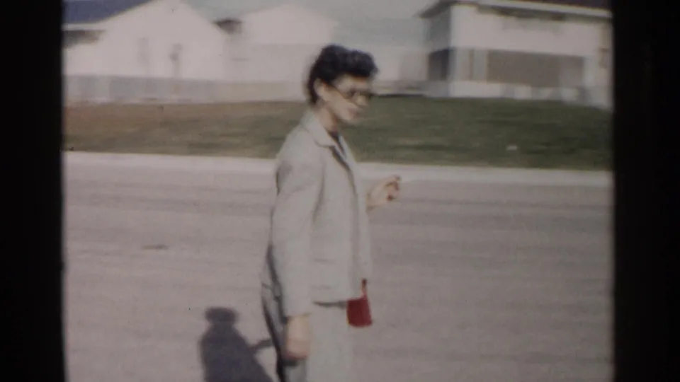 Closer 8mm view of the woman smiling in sunglasses and a pale suit as she walks along a Washington DC neighborhood street in 1962.
