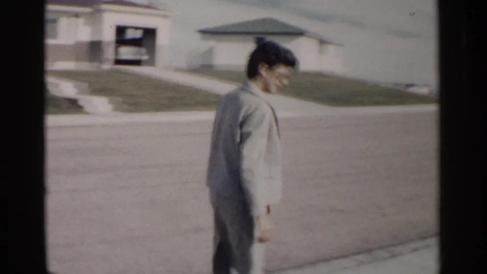 Profile view of the woman walking away on a sunlit Washington DC residential street, with modest houses and lawns visible in the 1962 footage.