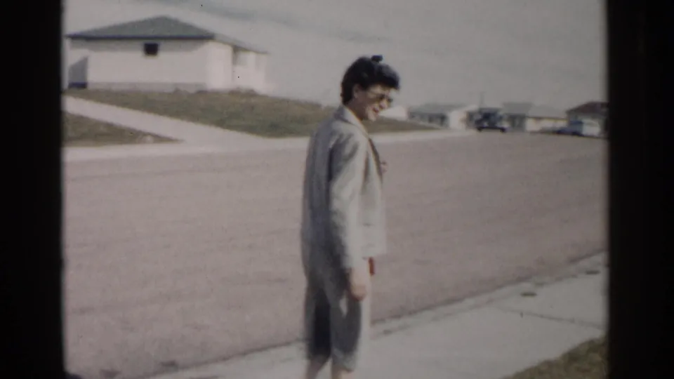 In 1962 Washington, DC footage, a woman in a grey suit walks along a sidewalk near single-story homes and parked cars.