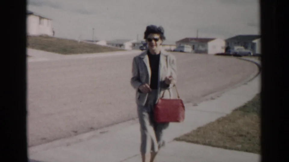 A woman in sunglasses and a grey suit carries a red handbag toward the camera on a Washington, DC residential sidewalk in 1962.