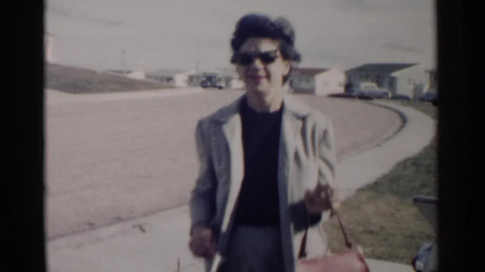 Closer 8mm frame of the woman in a grey suit and sunglasses walking with a red handbag on a sunny residential street.