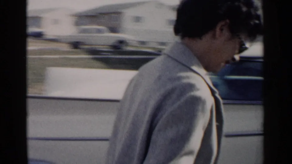 The woman passes the camera in profile beside a light-colored parked car, with residential houses blurred behind her.