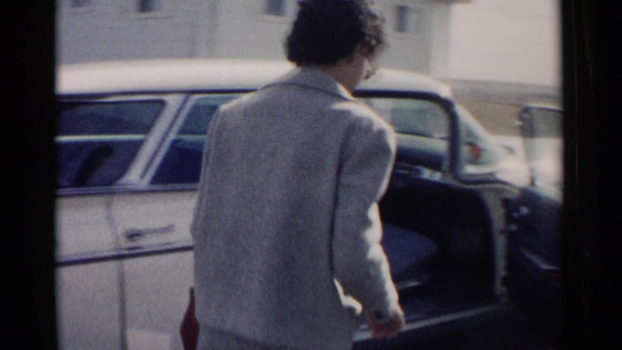 Woman in a light suit approaches an open rear door of a parked vintage car.