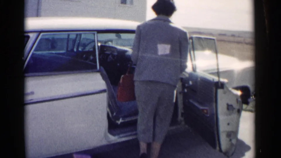 A woman in a light-colored outfit stands beside the open rear passenger door of a two-tone car in 1962 Washington, DC footage.