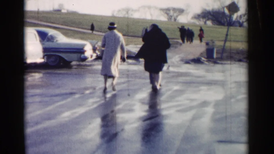 Two adults walk away on wet pavement in Washington, DC, with parked classic cars, distant pedestrians, and a grassy hill in view.