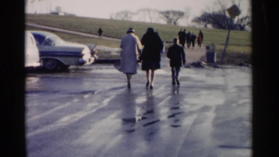 Adults and a child walk away along a wet Washington, DC street in 1962, with classic cars, distant pedestrians, and a grassy slope visible.