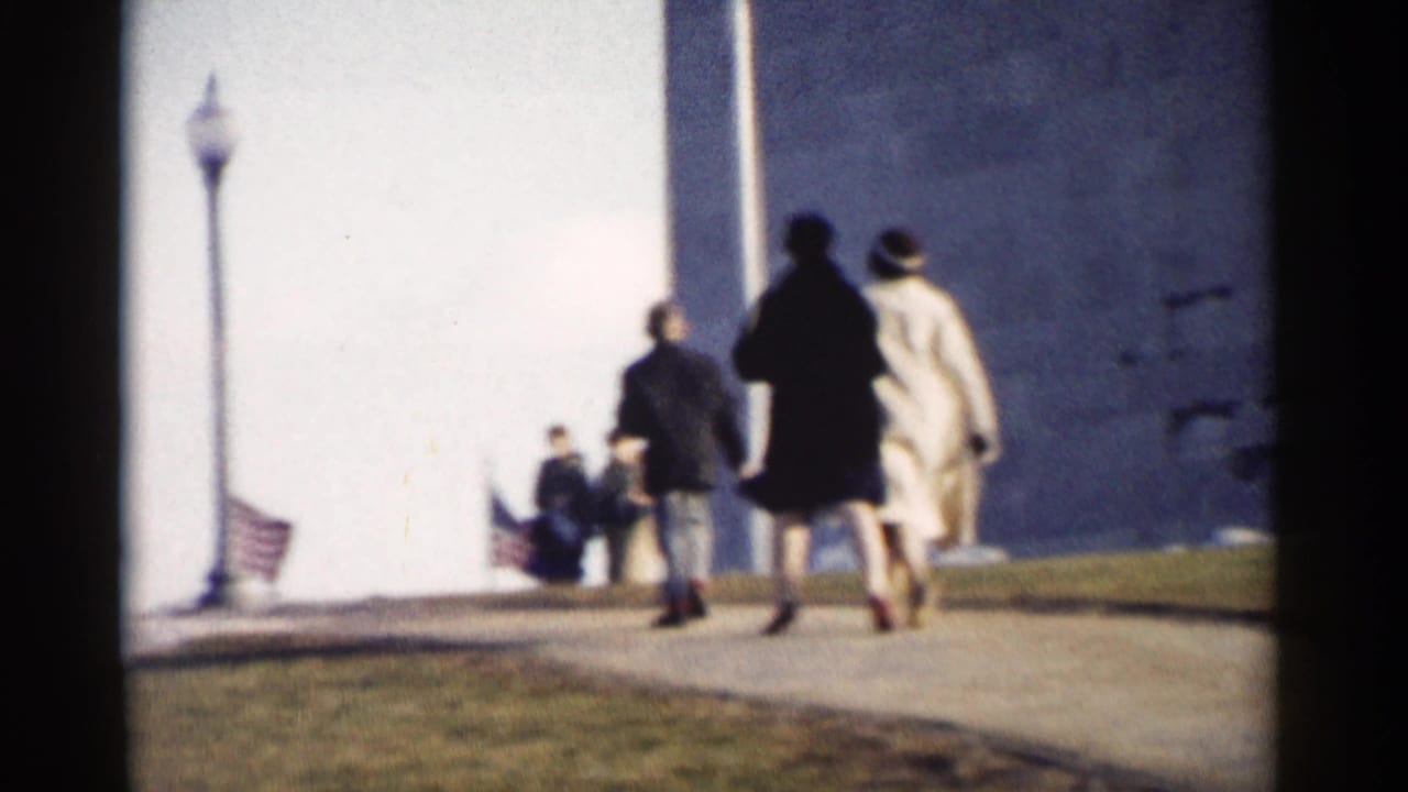 1962 Washington, DC home-movie frame showing visitors on a paved path near the U.S. Capitol exterior and small American flags.
