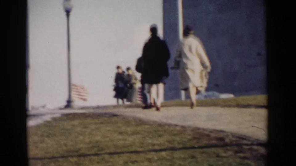 1962 Washington, DC frame of visitors walking past the Capitol exterior, with small American flags and a lamppost in view.