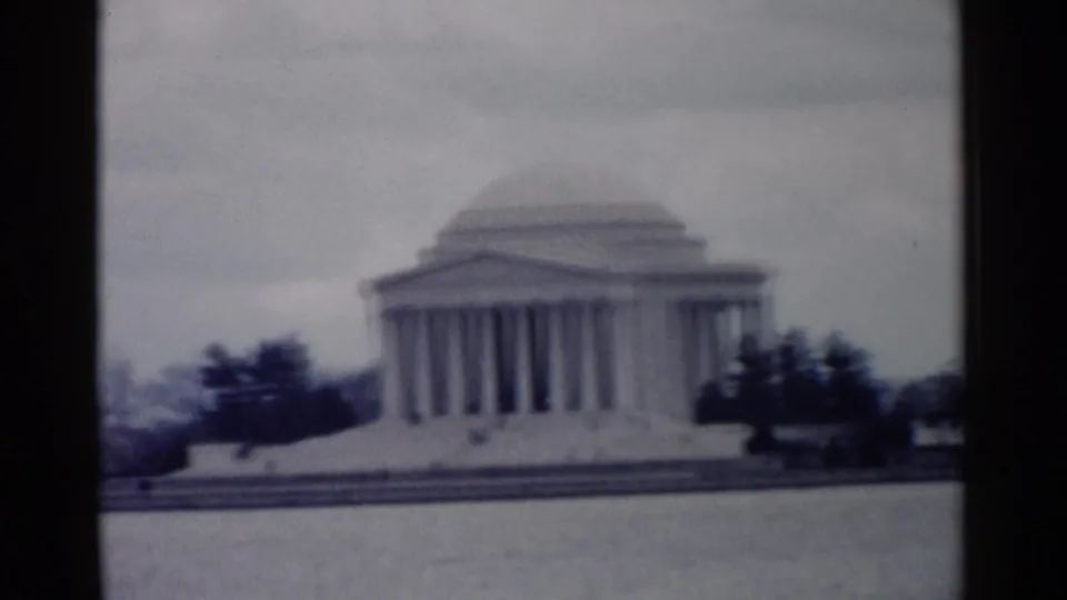 Soft 8mm frame of the Jefferson Memorial across the Tidal Basin, with water, trees, steps, and overcast sky visible.