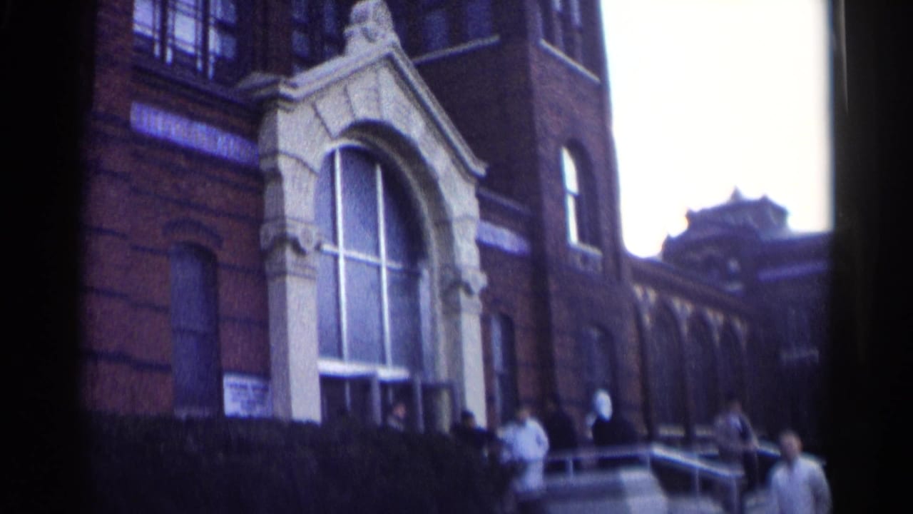 Brick school building with arched entrance, steps, and people near the doorway.