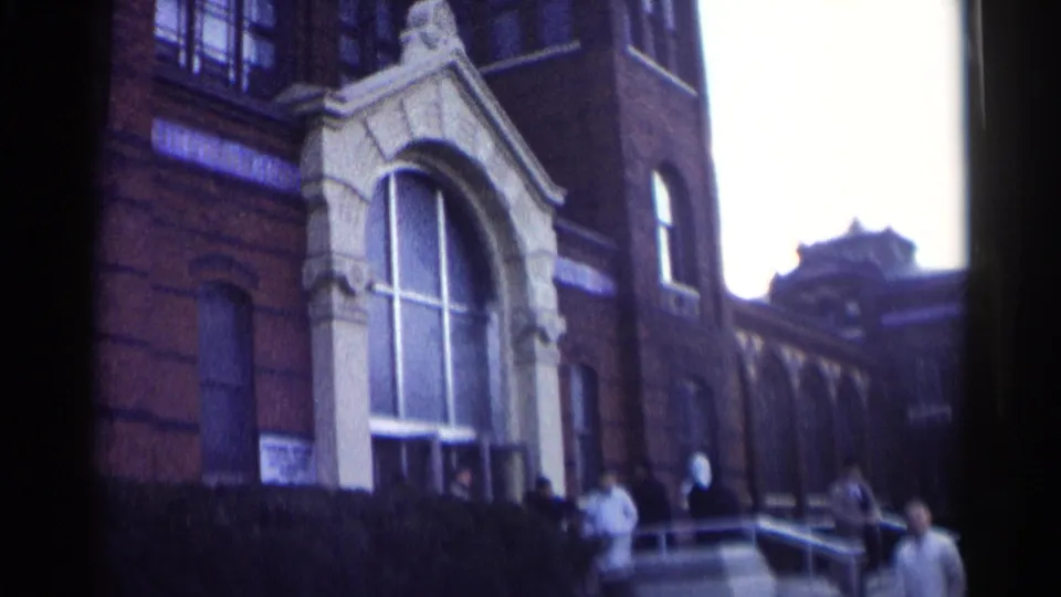 1962 Washington, DC 8mm frame showing a brick school exterior with an ornate arched entry and people near the steps.