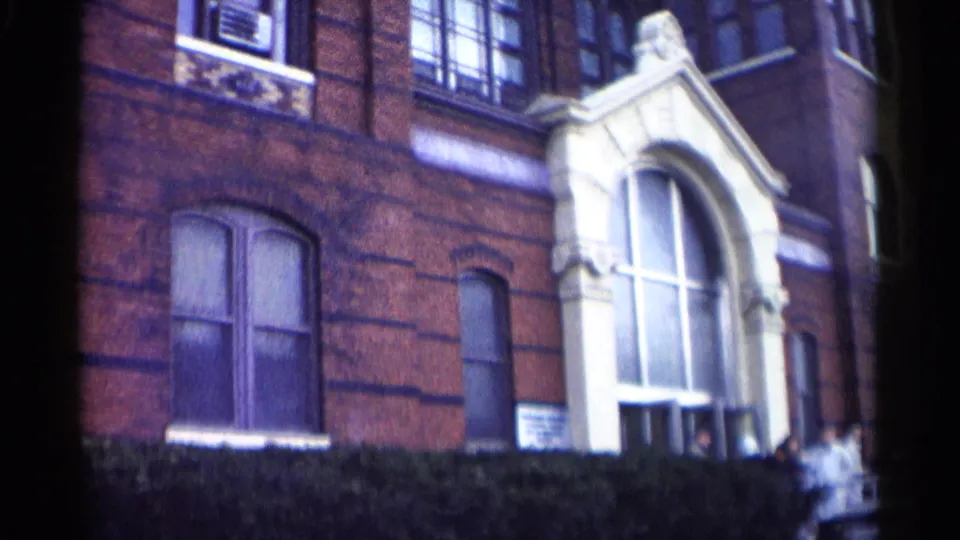 Wider 8mm view of the Washington, DC school entrance, with shrubbery below the facade and people near the right side.