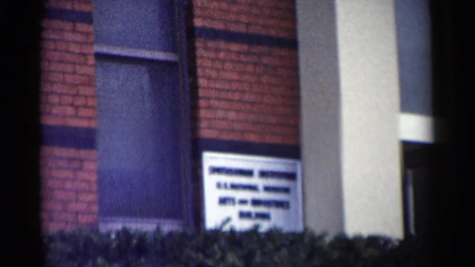 Close 1962 Washington, DC frame of a school sign mounted near a window on the brick exterior, with shrubs in front.