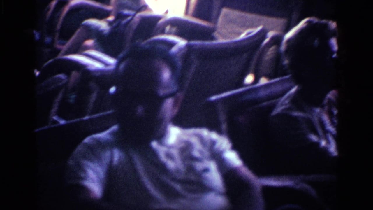 In 1963 Kentucky home-movie footage, two older adults sit in a dim living room, one man wearing glasses in the foreground.