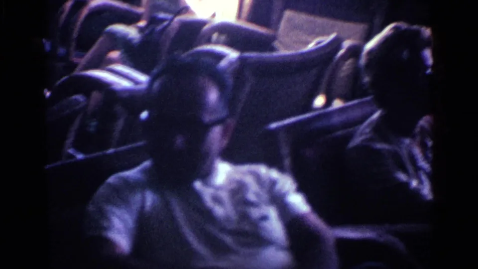 In 1963 Kentucky home-movie footage, two older adults sit in a dim living room, one man wearing glasses in the foreground.