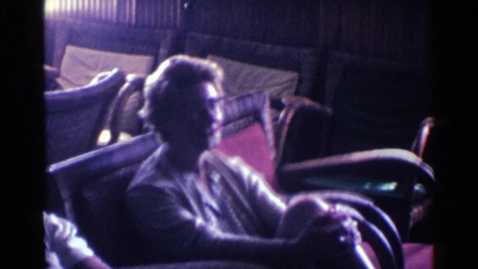 The camera angle shows an older adult seated near padded chairs, with dark paneling and soft 8mm color in the room.