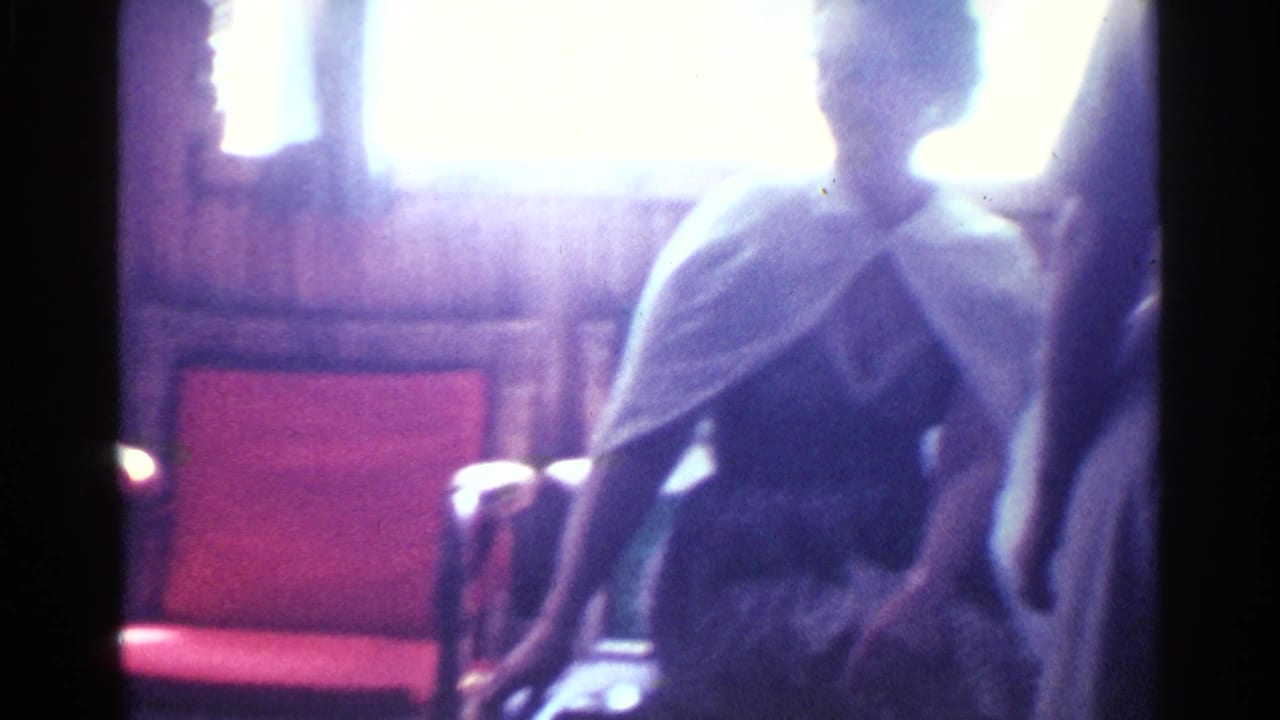 Backlit woman in a white shawl standing near a red chair in a wood-paneled room.