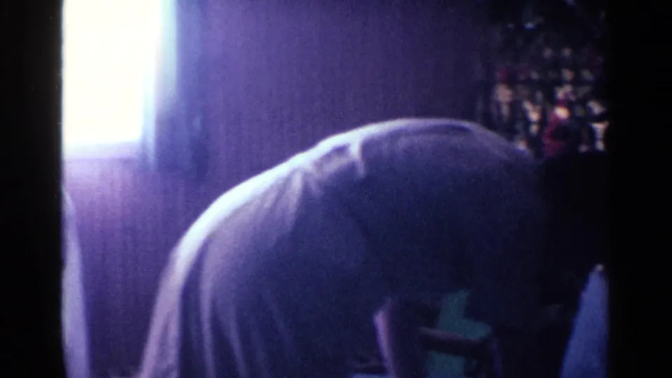 Dim 1963 Kentucky home-movie frame showing a person bending forward inside a wood-paneled private interior.