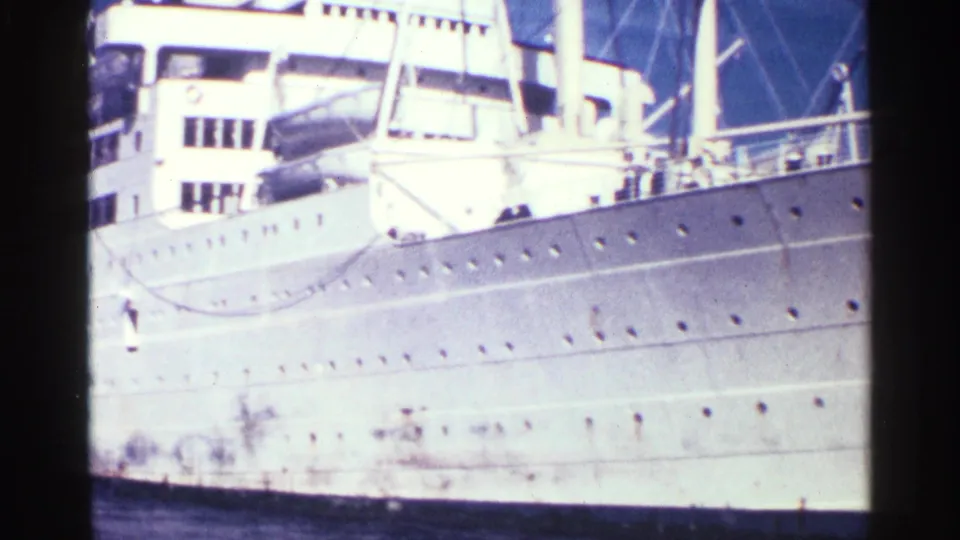 1963 home-movie frame of a white ocean liner’s side, showing portholes, deck railings, rigging, and sunlit superstructure.
