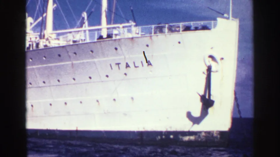 1963 home-movie frame showing the liner’s bow area, ITALIA lettering on the hull, and a large dark anchor against blue sky.