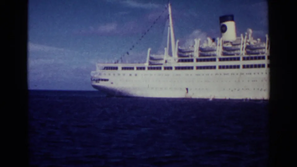 A wider 1963 8mm view shows the white Italia ocean liner in profile on calm blue water under a partly cloudy sky.
