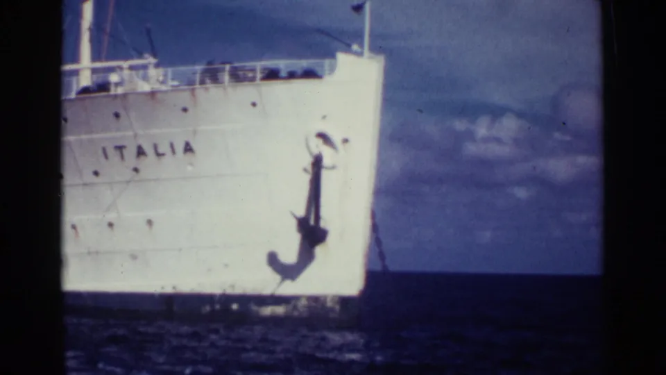 1963 Nassau 8mm frame showing a partial view of the Italia bow, with the hull name, anchor, ocean surface, and clouded blue sky in frame.