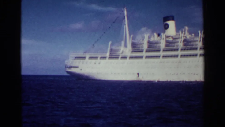 Wide 1963 Nassau home-movie view of the Italia at sea, showing the white passenger liner in profile with multiple decks, rigging, smokestack, blue sky, and dark water.