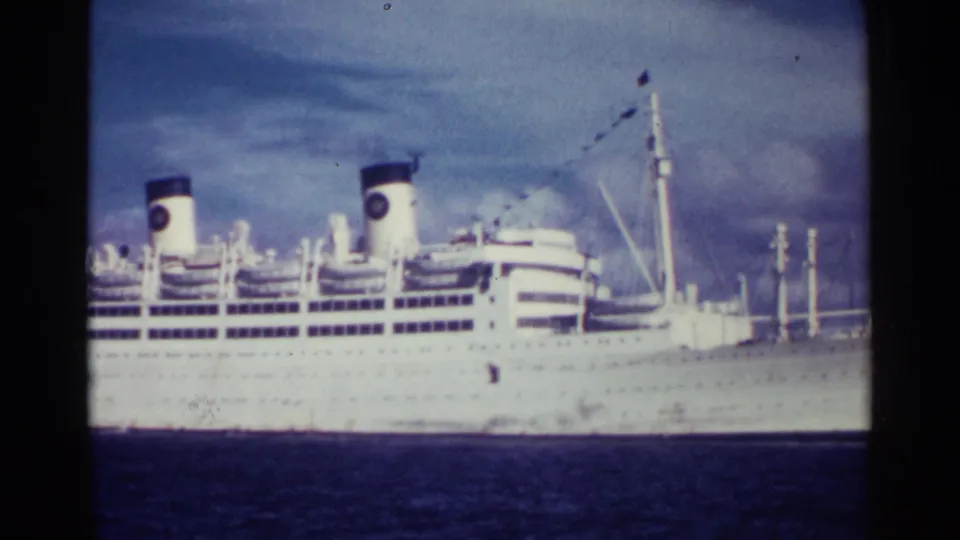 1963 Nassau frame showing the liner's side with paired funnels, deck lines, mast rigging, and stern area against blue sea and sky.