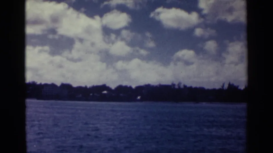 1963 Nassau shoreline view with calm blue water, low dark trees, small distant structures, and scattered clouds.