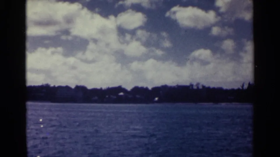 1963 Nassau coastal frame showing rippled water, a dark tree line, small pale shoreline buildings, and a bright clouded sky.