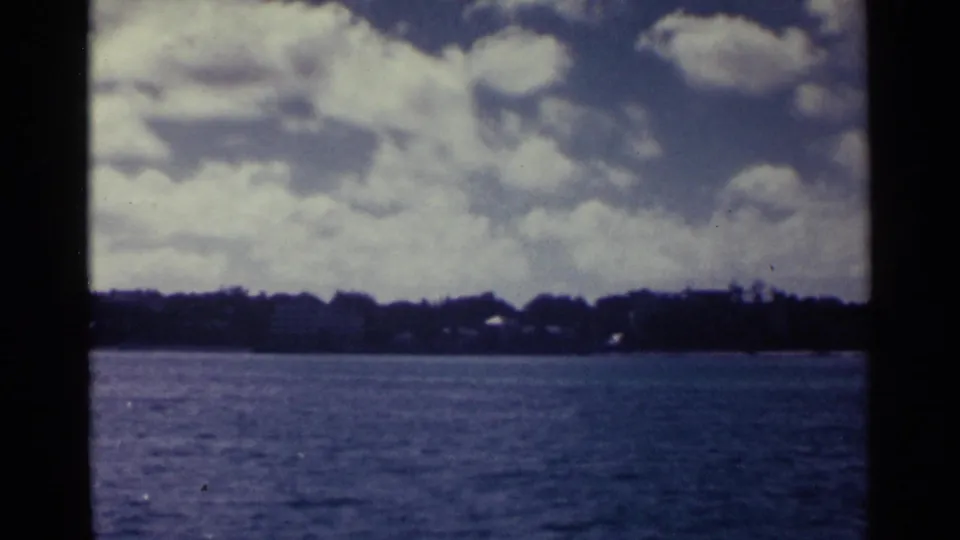 1963 Nassau home-movie view of rippled water, low shoreline, dark trees, small structures, and broad clouds overhead.