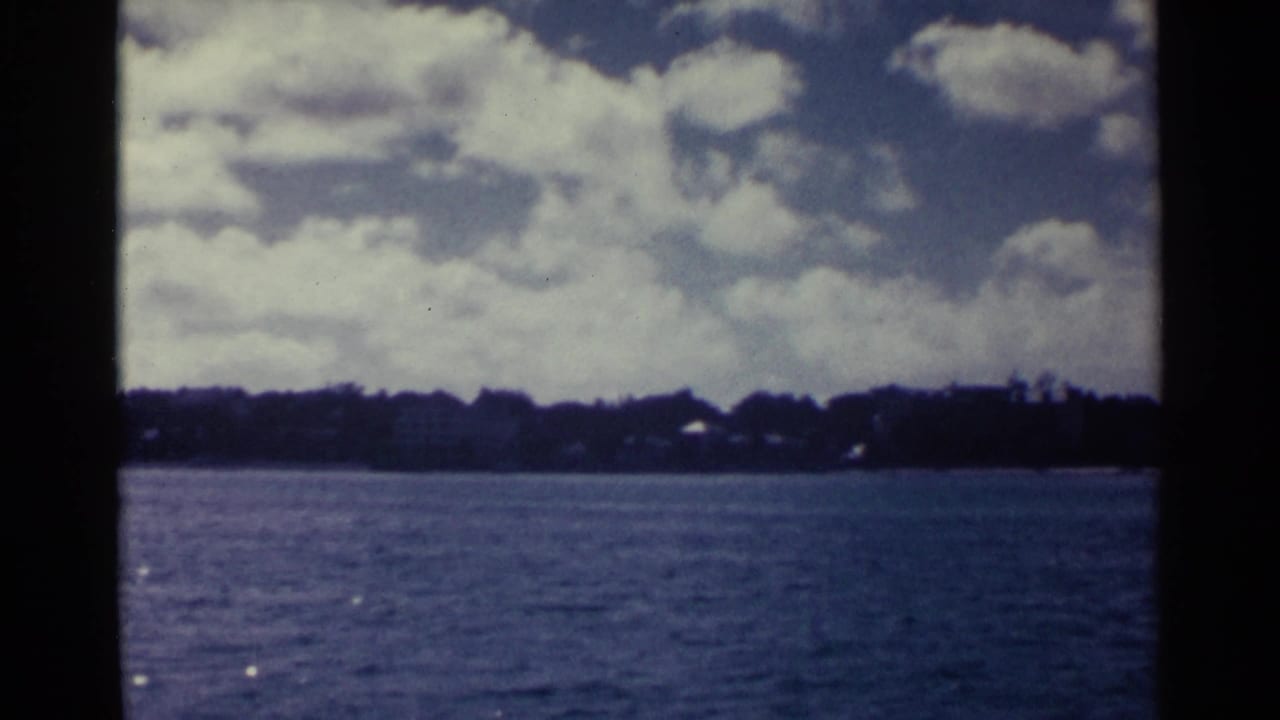 1963 Nassau waterfront view across blue water toward a low, tree-lined shoreline under a cloud-filled sky.