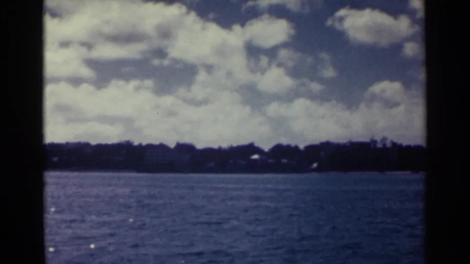 1963 Nassau waterfront view across blue water toward a low, tree-lined shoreline under a cloud-filled sky.
