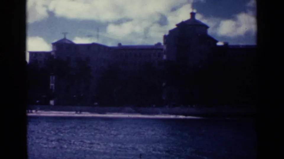 1963 Nassau home-movie frame showing a distant multi-story waterfront building beyond rippled water.